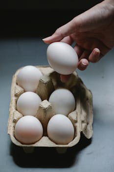 High angle of crop anonymous female taking raw chicken egg from cardboard container for cooking
